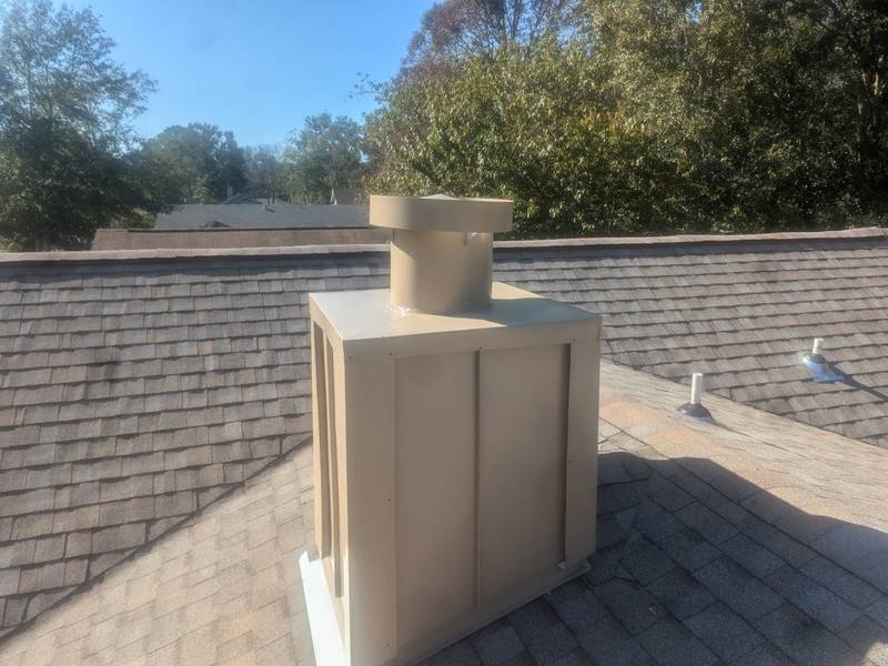 Tan metal-clad chimney chase with a round cap on a brown shingle roof, pipe vents and trees behind