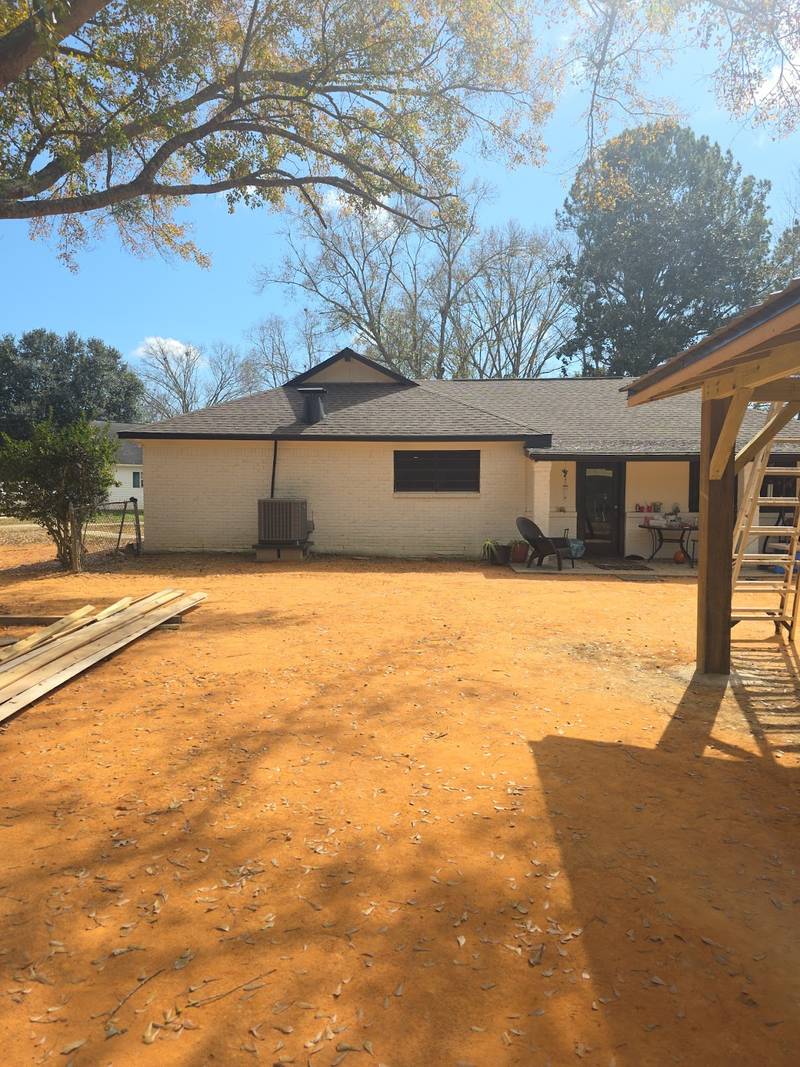 Back of a white-painted brick ranch house with a dark shingle roof, seen across an orange dirt yard under oak branches